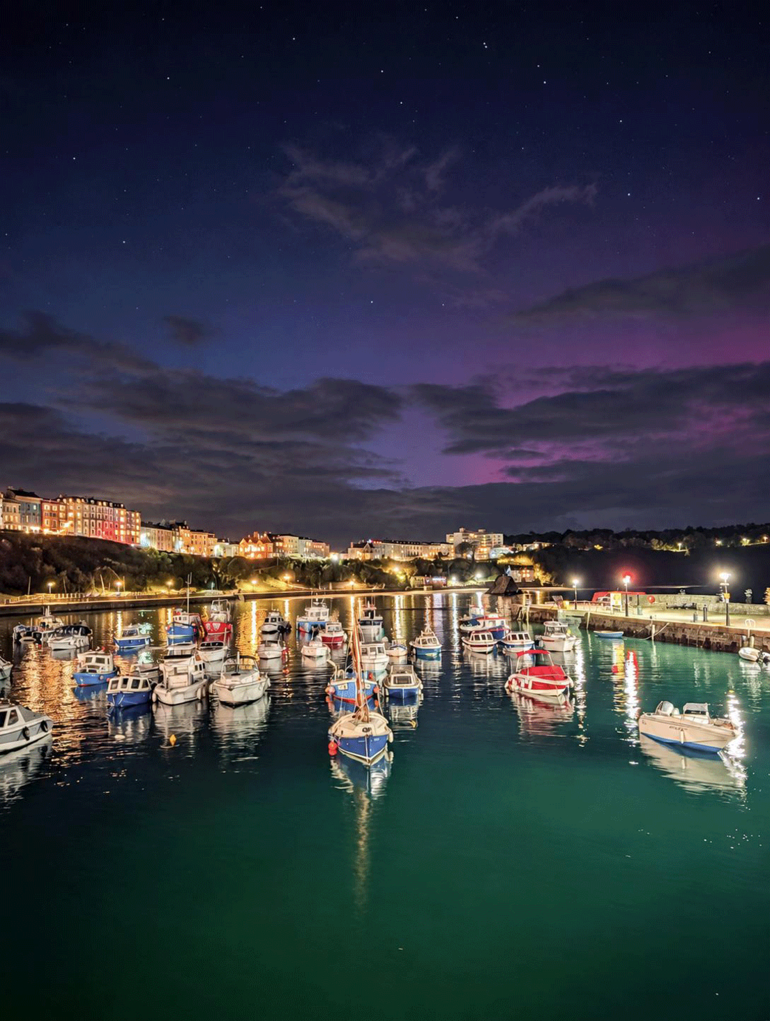 An image of the sky on 10 October during the evening. The purple hue of the Northern Lights can be seen above boats sitting on calm waters in Tenby harbour