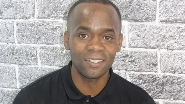Portrait of Anton Green wearing a black long-sleeved polo shirt, in front of a light grey brick-pattern wall. He faces the camera, with his head and shoulders visible in the frame. He is smiling.