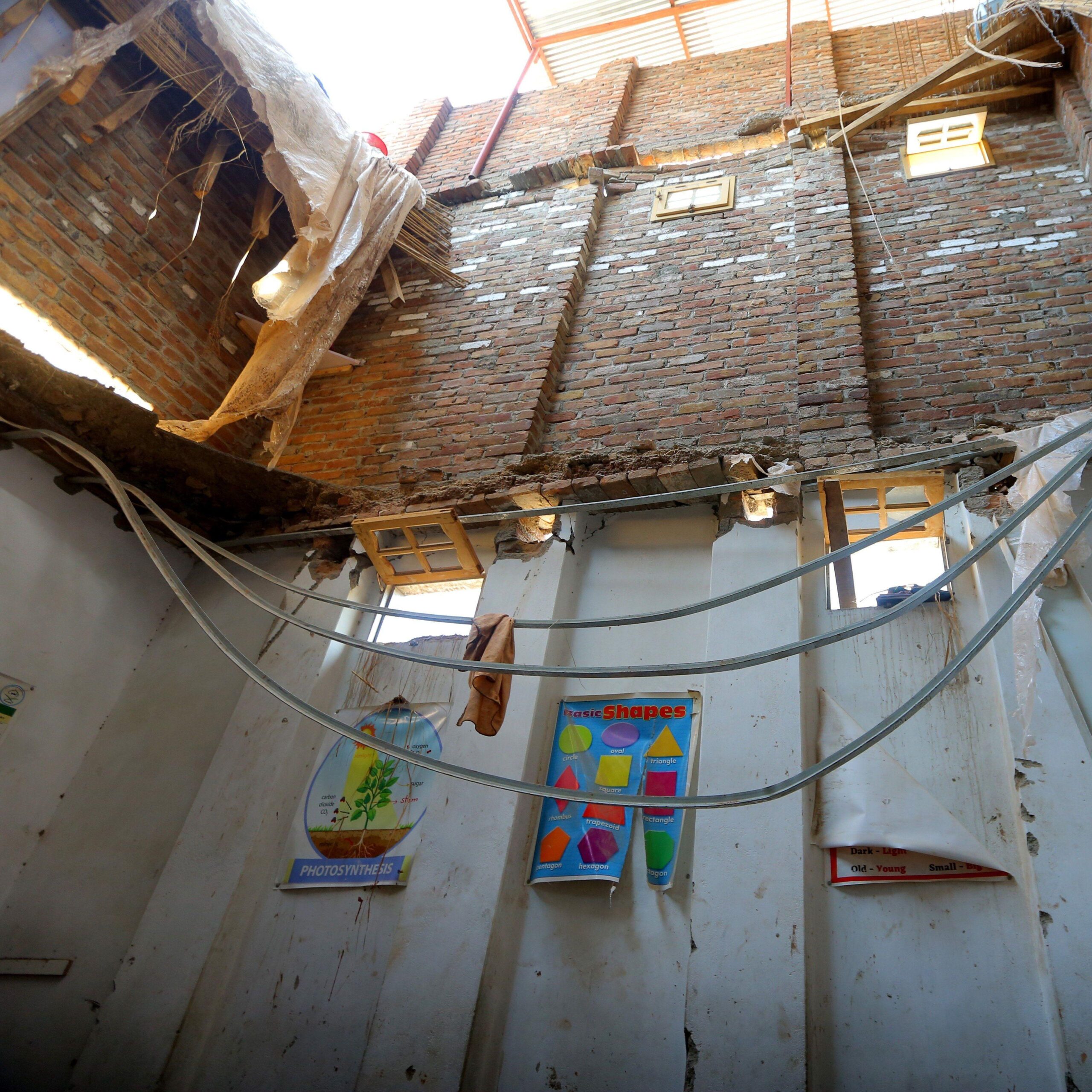 Interior of a damaged classroom with part of the roof collapsed, exposing brick walls and open sky above. Twisted metal ceiling supports hang across the room, while educational posters, including one on photosynthesis and another showing basic shapes, remain attached to stained white walls.