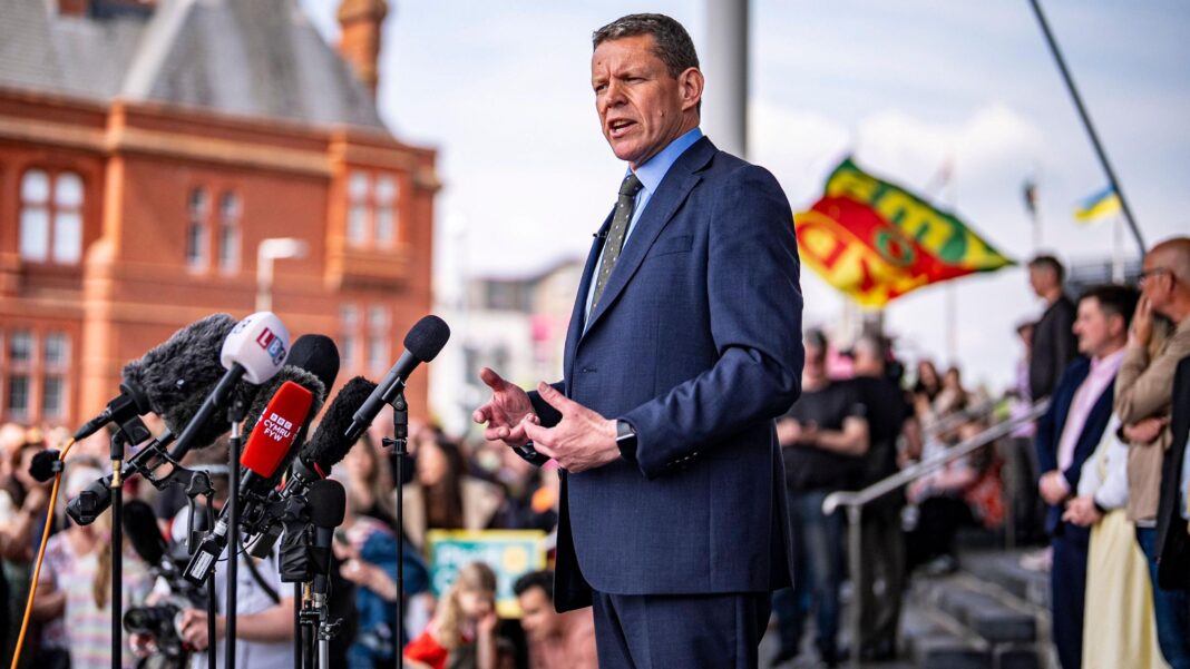 Rhun ap Iorwerth in a dark blue suit in front of microphones at Cardiff Bay in front of people gathered.