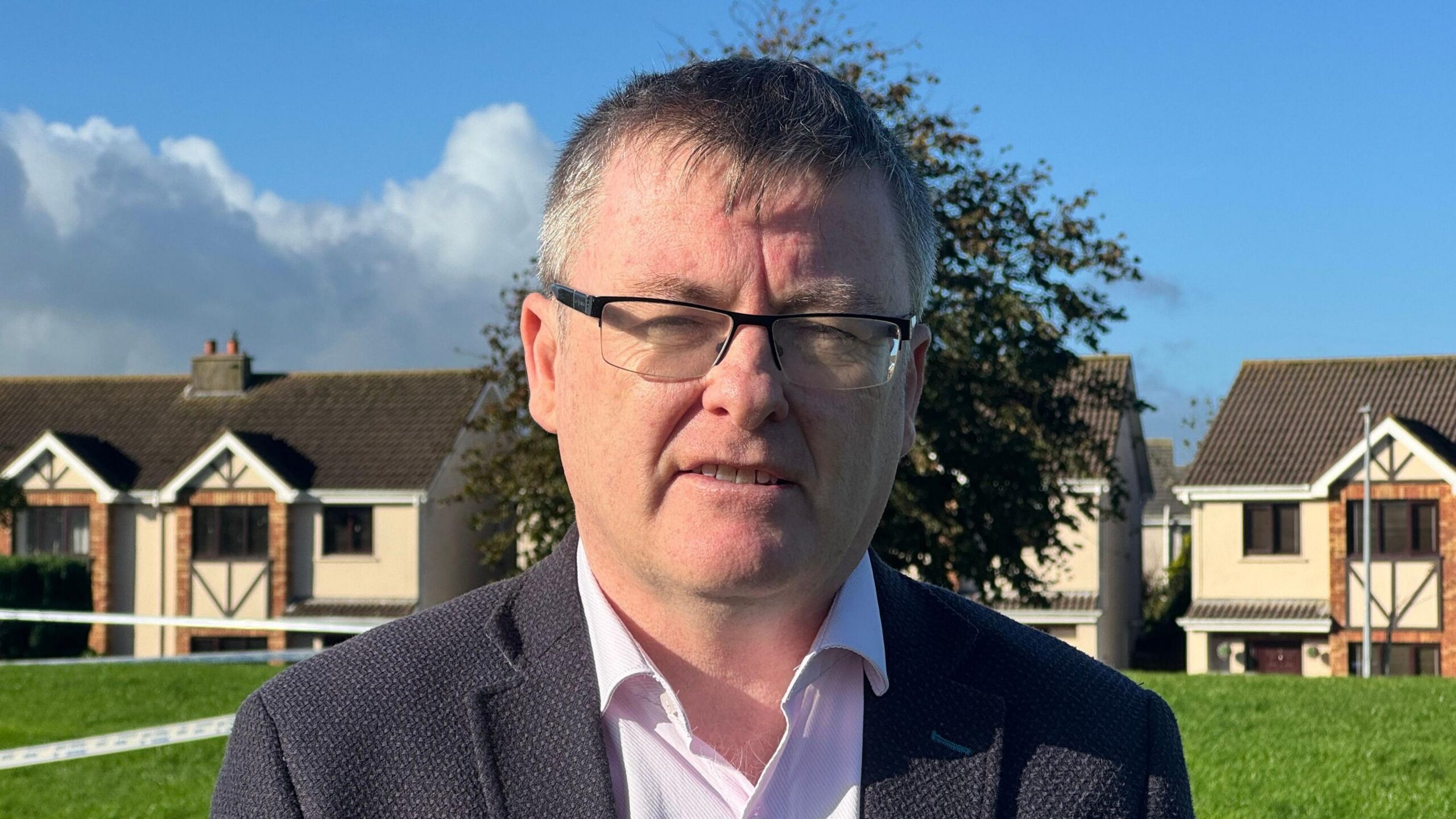 David Cullinane, a man with short, grey hair and glasses, looks at the camera while standing at the scene of a house fire.  He is wearing a navy suit jacket and an open-necked white or pale pink shirt.  There is a row of semi-detached houses in the background, some of which are cordoned off by white and blue tape.