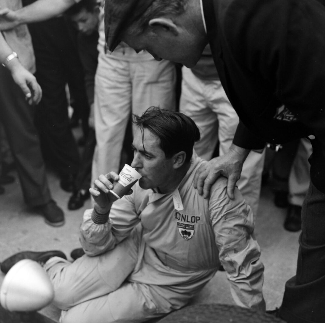 An exhausted-looking Jack Brabham takes a drink from a cup shortly after pushing his car over the line and winning the drivers' championship at the 1959 US Grand Prix at Sebring