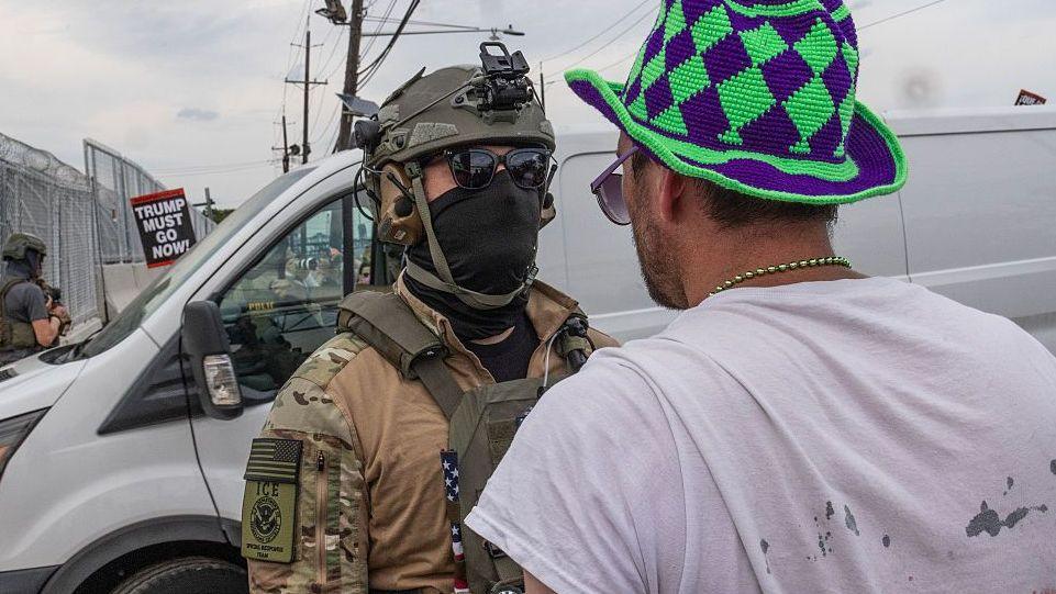 A US Immigration and Customs Enforcement police uniform confronts a man wearing a white shirt and a green and blue hat. A vehicle is visible behind them