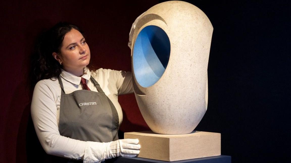 A woman with brown hair wearing an apron labelled Christie's is wearing white gloves and handling  the pale creamy brown sculpture which has a blue painted hollow centre. It is based on an ovoid shape and is balanced delicately on its narrowest point.