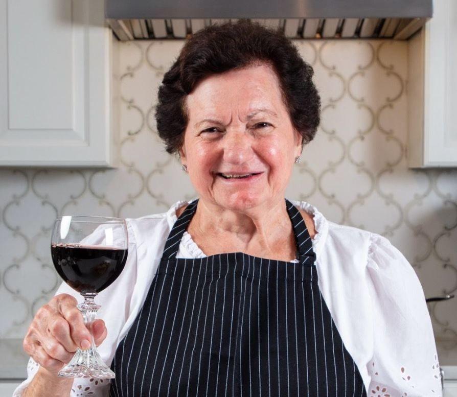 Nonna Gracie wearing a black apron with white pinstripes over a white top, and holding a glass of red wine