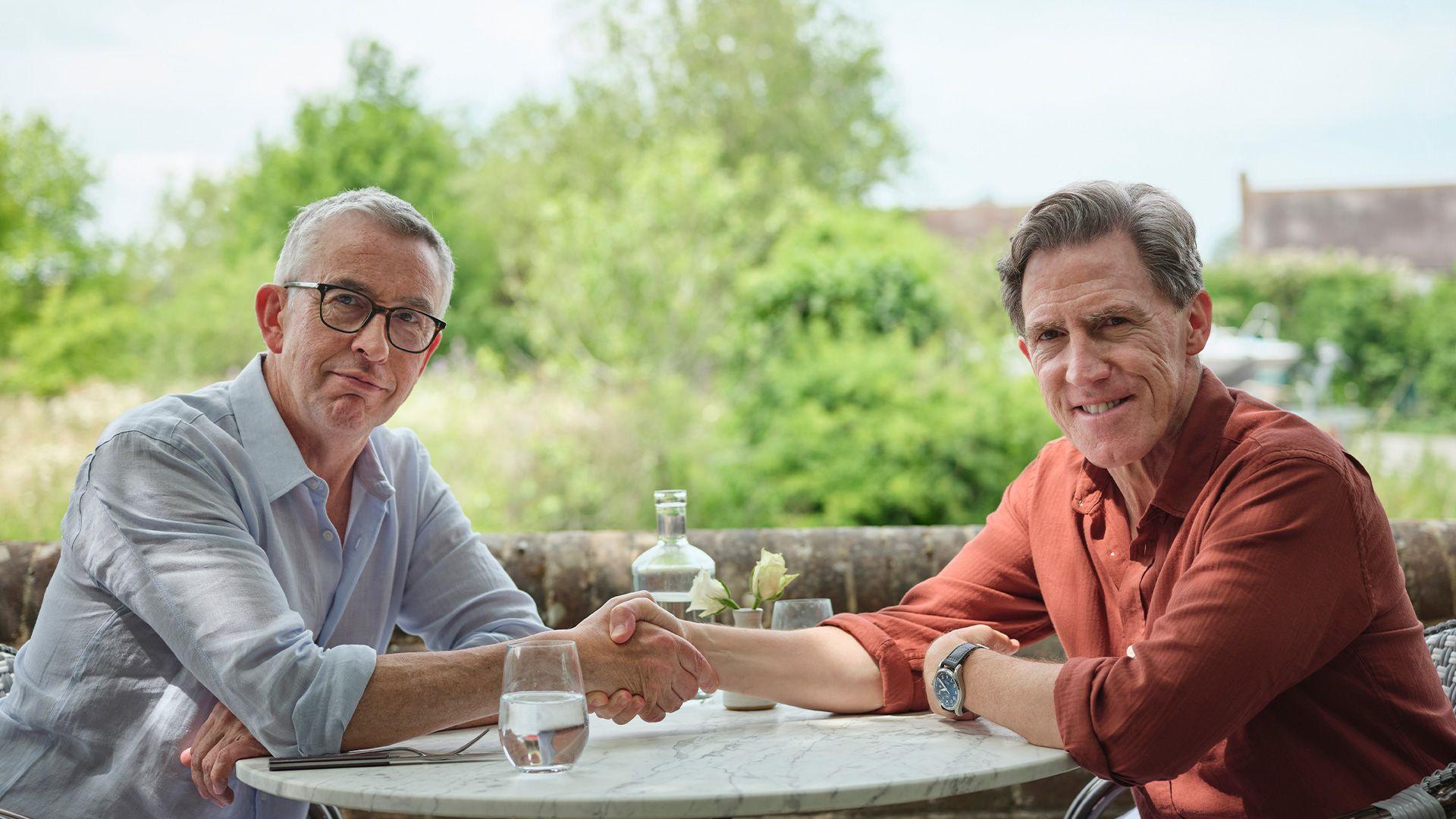 Steve Coogan and Rob Brydon sit at an outdoor café table, smiling and shaking hands. Steve wears a light blue shirt and black glasses. Rob wears a red shirt. They both have short grey hair and are seated facing the camera with glasses of water and a bottle between them, with green countryside and buildings blurred in the background.