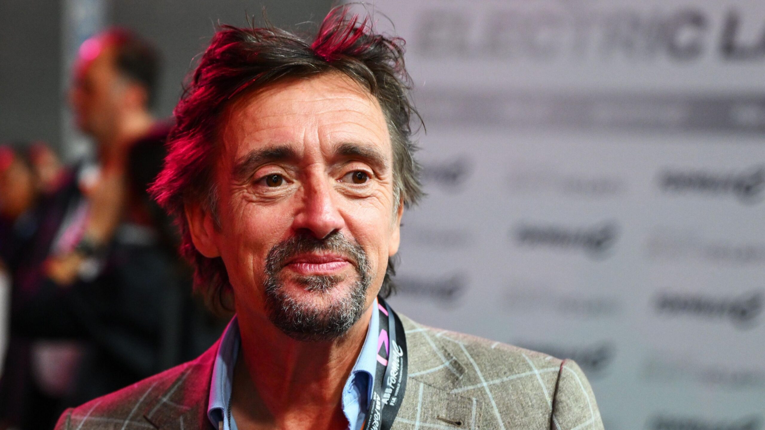 A head and shoulders shot of Richard Hammond. He has dark hair and a goatee and is wearing a grey chequered blazer and blue shirt, with a black lanyard just visible.