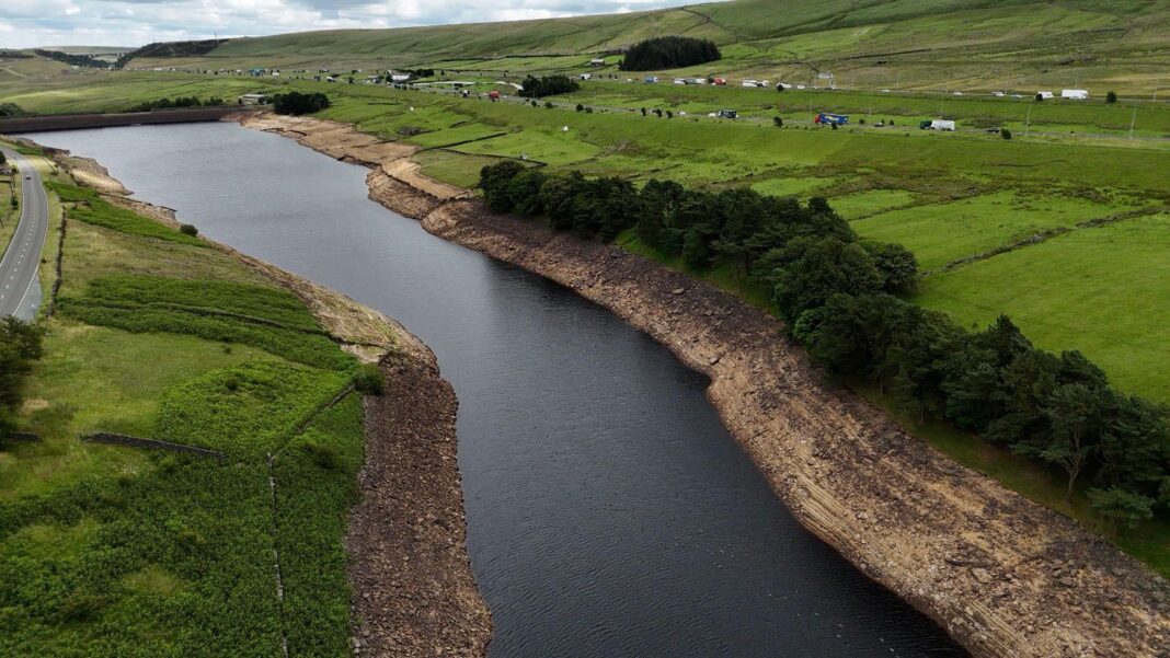 Drought declared in Yorkshire after dry weather Aerial view of a reservoir with visibly low water levels, exposing wide, dry, rocky banks. Surrounding the reservoir are green fields, scattered trees, and a road running parallel to the water.