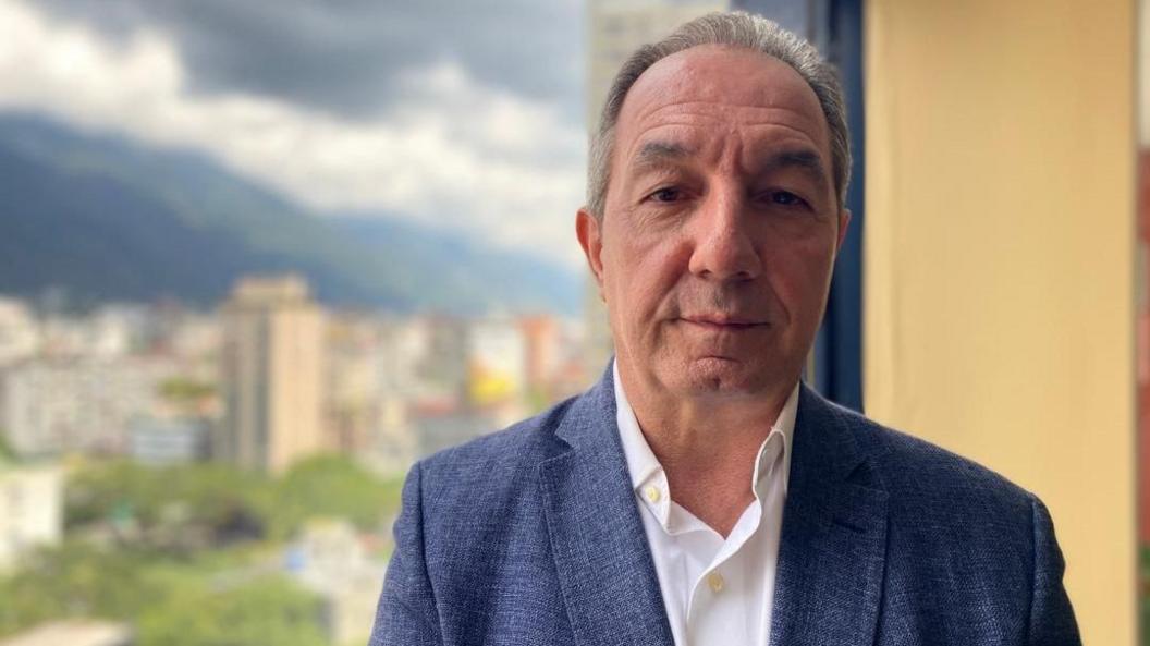 Political analyst Beningno Alarcon, a middle-aged man, is wearing a blue suit over a white shirt. The blurred backdrop of a city's skyline is also in view.