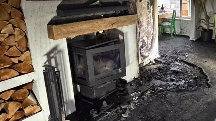 A log burner, in a room, with smoke and fire damage. Stacks of wood are to the left. The carpet is burnt and black. A table and green chair can be seen at the back of the room. A wood plinth is above the log burner, which is black.